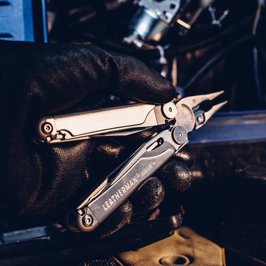 Translation missing: en. A person wearing black gloves holding a Leatherman Wave+, showcasing it's pliers and tools inside the handle, getting ready to work on a vehicle