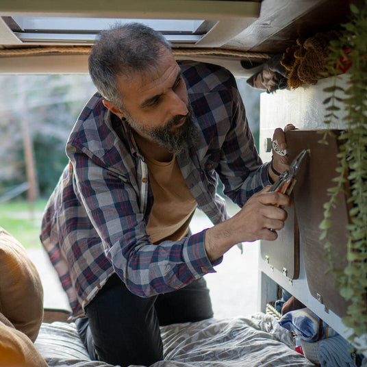 Translation missing: fr.Person using a Leatherman Signal multi-tool to make repairs inside a camper van, kneeling on a bed while gripping the tool to tighten a fastener on a cabinet panel