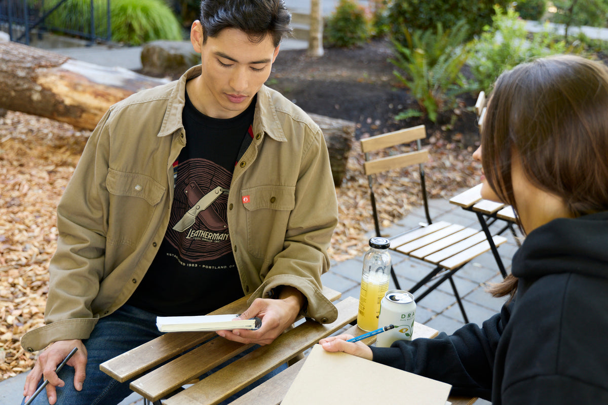 Translation missing: it.A man sitting at a table wearing a black Leatherman Blade & Grain tee with a woman wearing a black hoodie, both equipped with a notebook and pencil