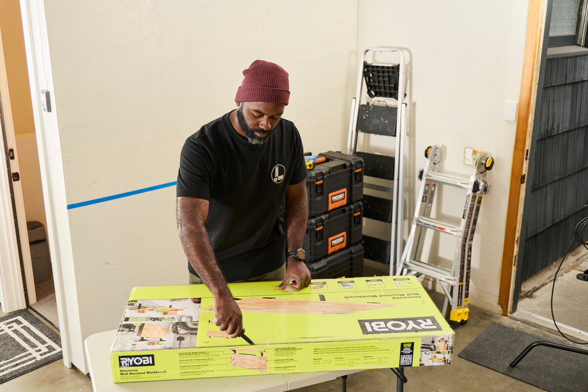 Translation missing: de.A person wearing a black Leatherman Classic Tee using the blade on a Leatherman multi-tool to open a large box that is laying on top of a folding table