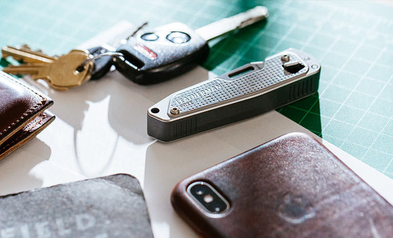 Translation missing: de.Brown Leatherman Wallet, car keys, Leatherman T2, gray note pad, and Iphone on top of white piece of paper placed on green grid table