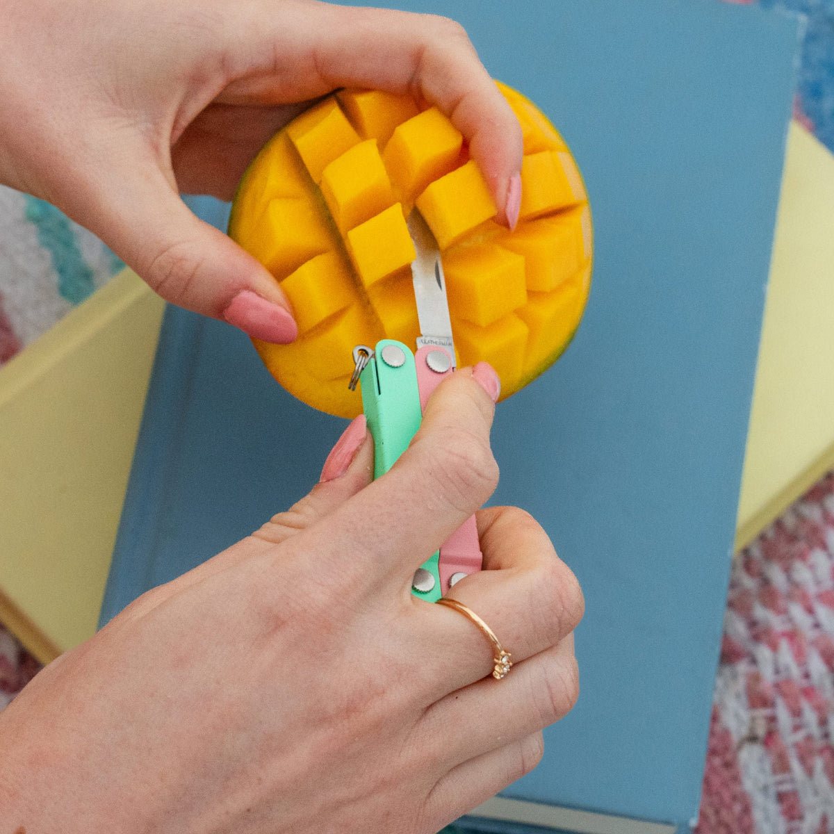 Translation missing: es.Hands using a Leatherman Micra® in Bayside color to cut a mango on top of a stack of books