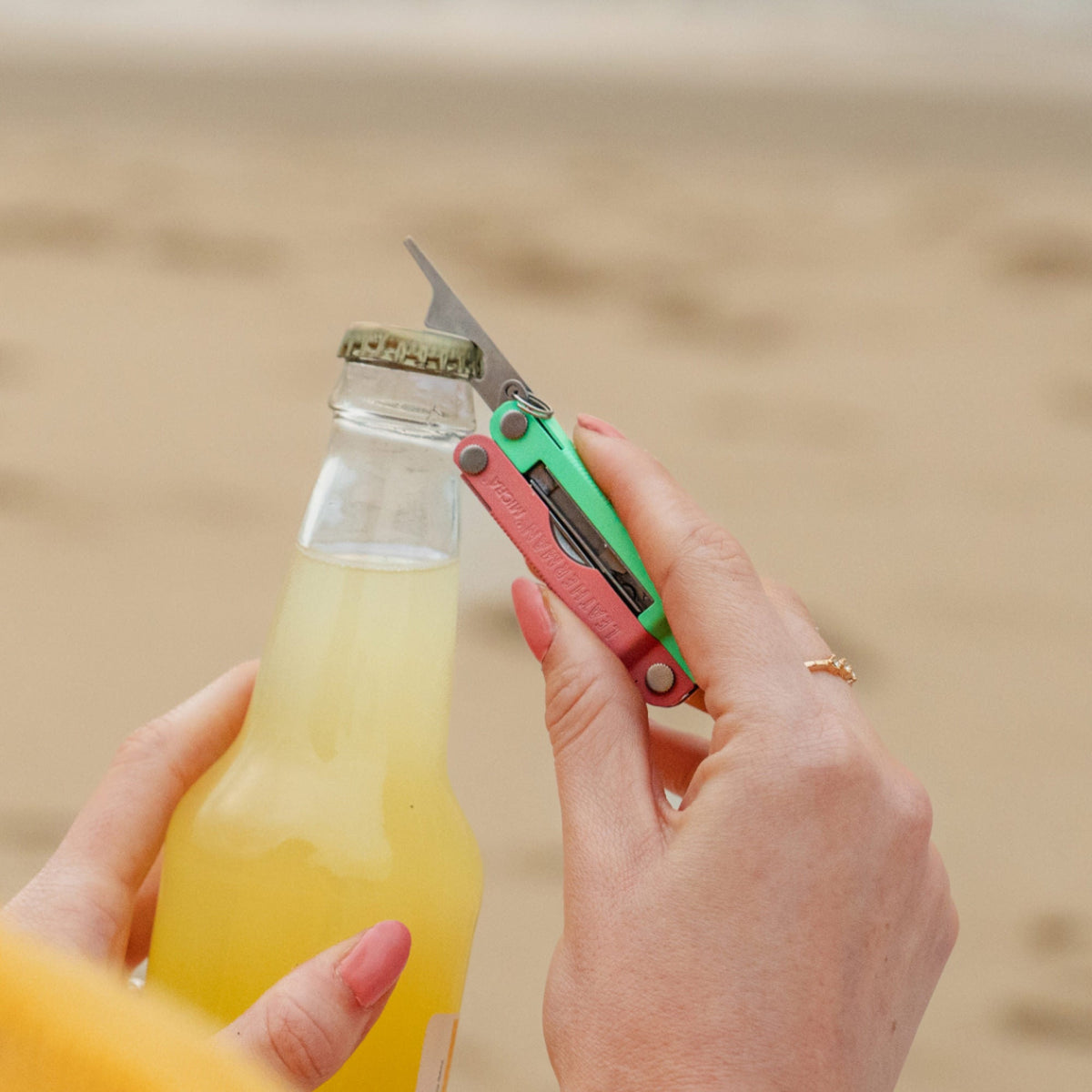 Translation missing: es.Person using the bottle opener on a Leatherman Micra® in Bayside color to open a bottle on the beach