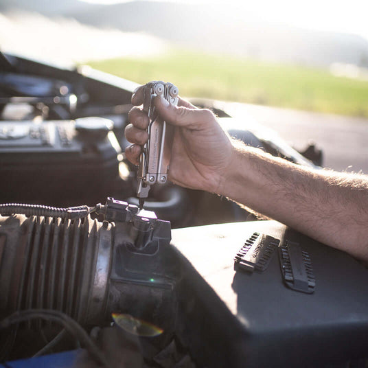 Translation missing: de.Person using a Leatherman Surge® multitool on a car engine in a sunny outdoor setting