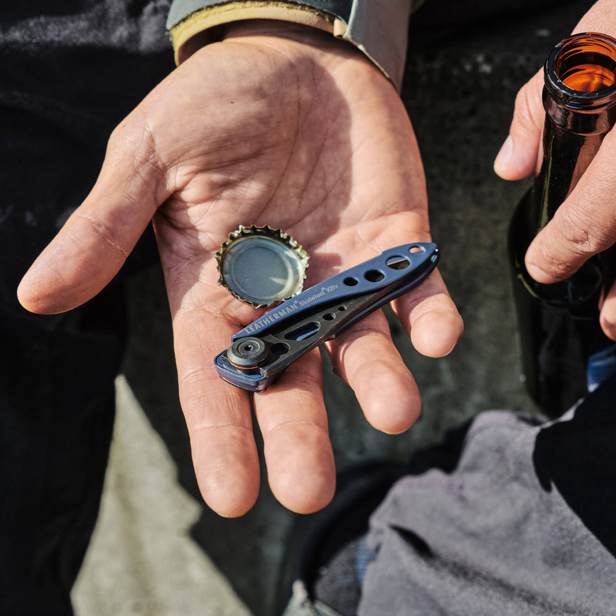 Translation missing: fr.Hand holding a blue Skeletool KBX and bottle cap next to a person gripping an open glass bottle