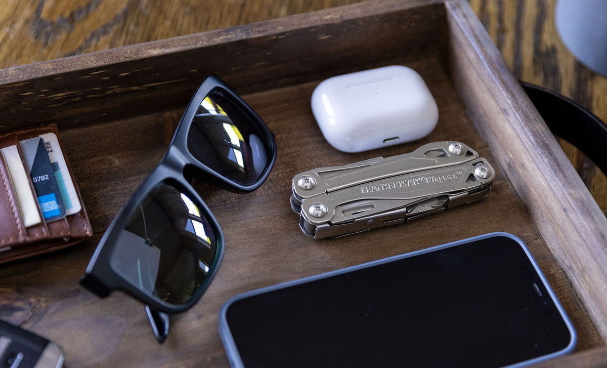 Translation missing: es.Stainless steel Leatherman Wingman® multi-tool beside sunglasses, smartphone, and AirPod case in a wooden tray