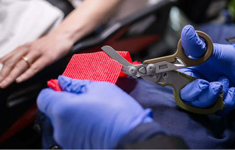 Translation missing: de.A person wearing blue gloves using the shears on a Woodland colored Leatherman Raptor Rescue to cut a piece of red medical fabric