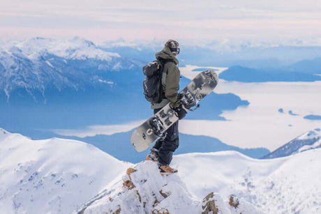Translation missing: de.A person with a backpack on standing on the top of a snowy mountain holding a snowboard