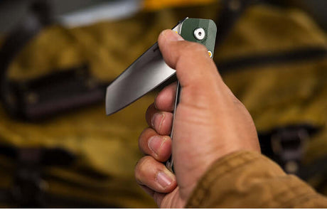 Translation missing: de.Close-up of a person holding a partially open Leatherman folding knife with a green handle, positioned in front of a yellow canvas bag with leather straps
