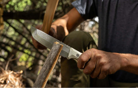 Translation missing: de.A person using a Leatherman knife with a white handle to split a piece of wood outdoors. The scene is set in a wooded area with dappled sunlight, and the individual is performing a batoning technique with a secondary stick to strike the back of the blade