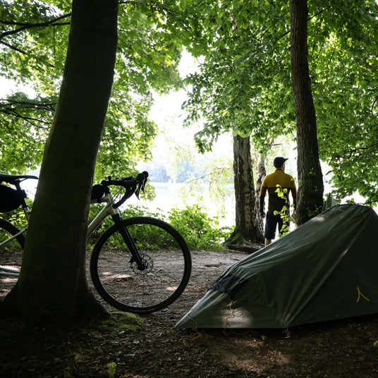 Translation missing: es.Cyclist standing beside a tent and bicycle in a forest clearing near a lake