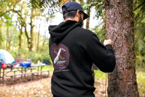 Translation missing: de.A person wearing a black Leatherman Blade &amp; Grain hoodie standing next to a tree in a forest campsite. The hoodie’s back graphic shows a pocketknife illustration with “Leatherman Tool Group” text