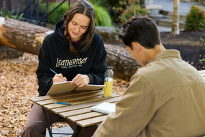 Translation missing: de.Two people sitting at a table outside with notebooks and pencils. One is wearing a black Leatherman Blade &amp; Grain Hoodie with 'Leatherman Tool Group Inc.' on the front