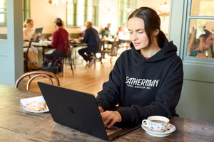 Translation missing: de.A woman sitting indoors working on a computer while wearing a black Leatherman Blade &amp; Grain hoodie with a pastry and coffee on either side of the laptop