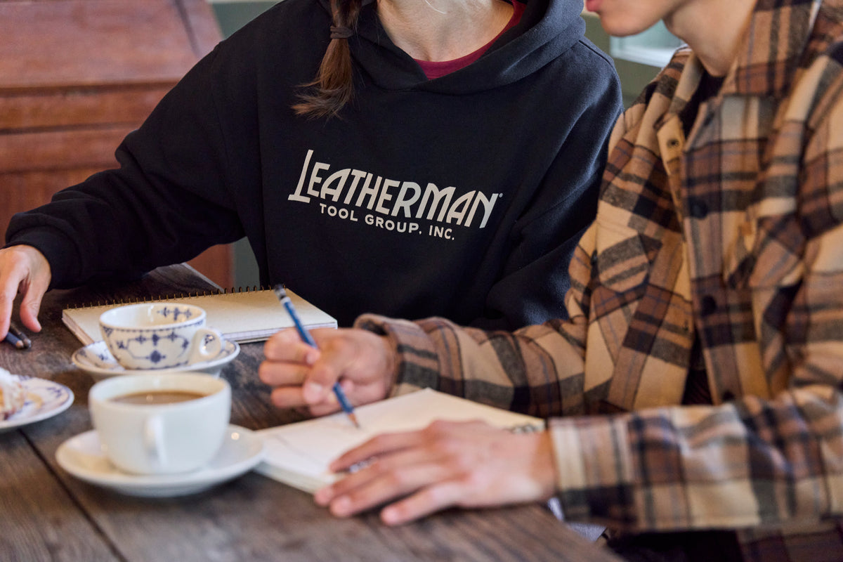 Translation missing: de.Two people sitting at a table with coffee, pastries and notebooks. One is wearing a black Leatherman Blade &amp; Grain hoodie with 'Leatherman Tool Group Inc.' on the front