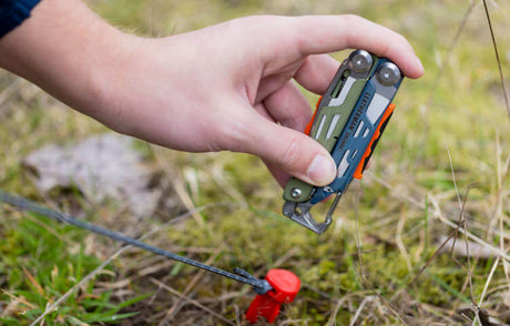 Translation missing: de.A person uses Leatherman Signal to hammer tent stake in patch of grass