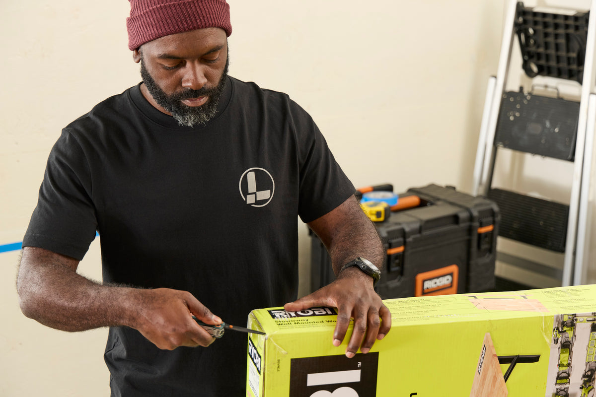 Translation missing: de.A person wearing a black Leatherman Classic Tee using the blade on a Wave Alpha in Cascadia color to open a large box in a workshop while standing near a ladder and tool case