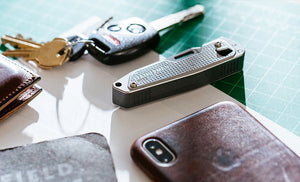 Translation missing: de.Brown Leatherman Wallet, car keys, Leatherman T2, gray note pad, and Iphone on top of white piece of paper placed on green grid table