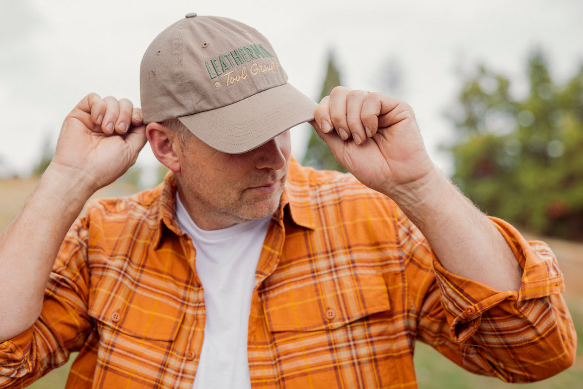 Translation missing: fr.A person wearing a Leatherman Dad Hat in Driftwood color adjusting the brim while outdoors, dressed in an orange plaid shirt