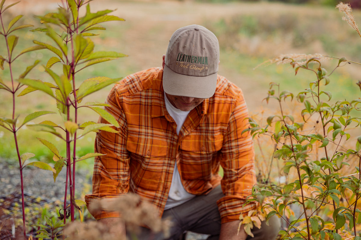 Translation missing: fr.A person wearing a driftwood colored Leatherman Dad Hat tending to plants outdoors while dressed in an orange plaid shirt