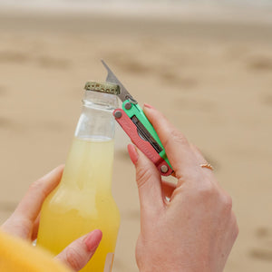 Translation missing: es.Person using the bottle opener on a Leatherman Micra® in Bayside color to open a bottle on the beach