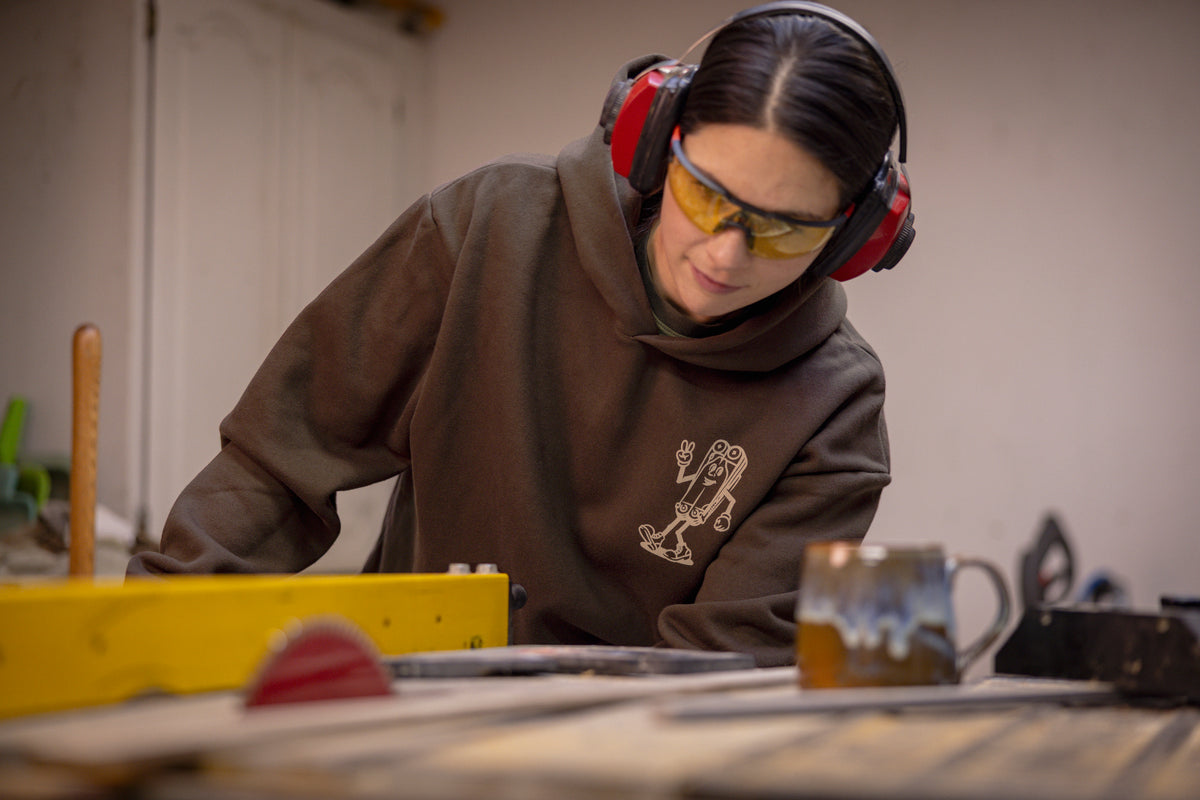 Translation missing: de.A person wearing a brown Leatherman Mr. Wave hoodie operating a table saw in a workshop, wearing safety glasses and earmuffs for protection