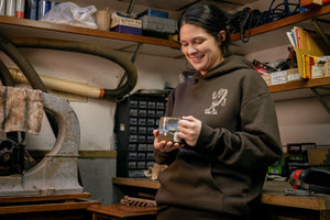 Translation missing: de.A person wearing a brown Leatherman Mr. Wave hoodie standing in a workshop holding a ceramic mug, smiling while surrounded by tools and equipment