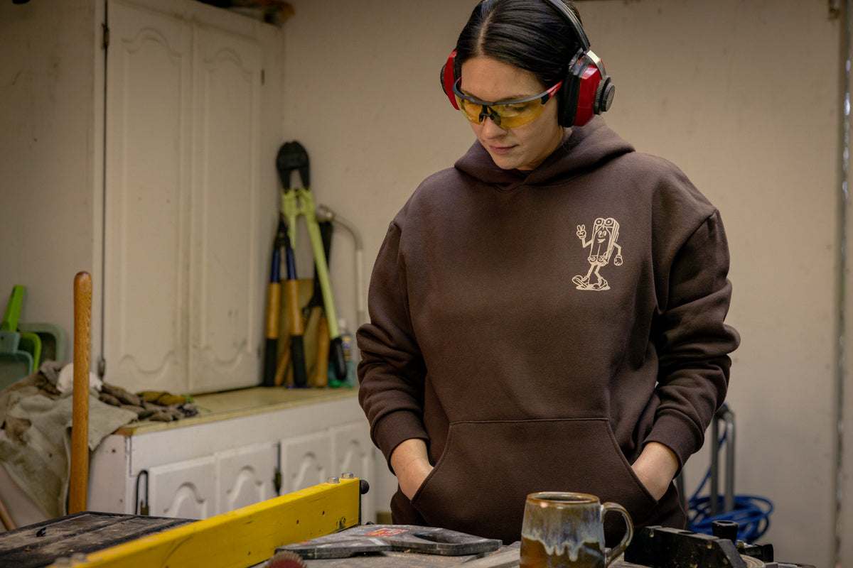 Translation missing: de.A person wearing a brown Leatherman Mr. Wave hoodie standing in a workshop with safety glasses and ear protection, looking down at a workbench with tools and a ceramic mug