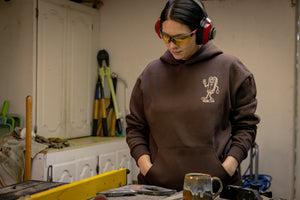 Translation missing: de.A person wearing a brown Leatherman Mr. Wave hoodie standing in a workshop with safety glasses and ear protection, looking down at a workbench with tools and a ceramic mug