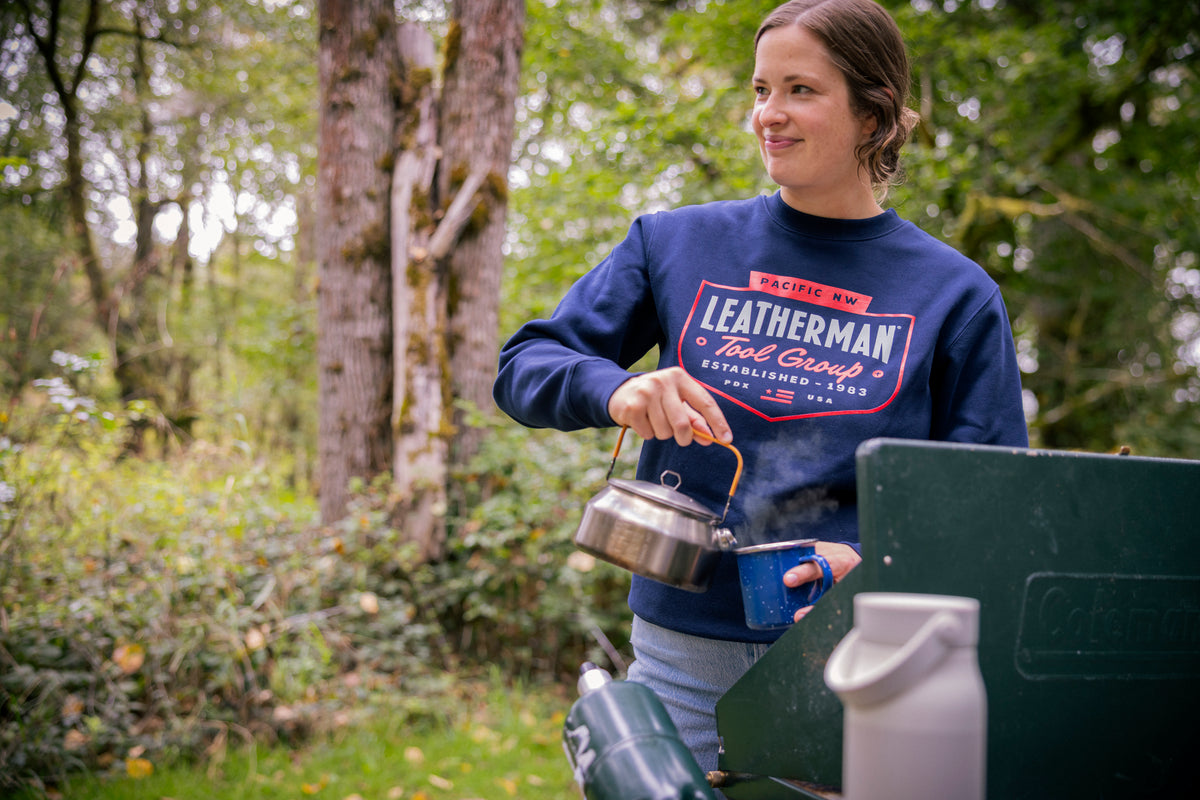 Translation missing: es.A person wearing a navy blue Leatherman PNW Crewneck sweatshirt pouring hot water from a kettle into a mug at a campsite surrounded by trees