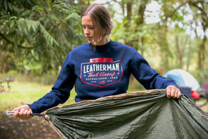 Translation missing: es.A person wearing a navy blue Leatherman PNW Crewneck sweatshirt setting up a hammock at a forest campsite with a tent in the background
