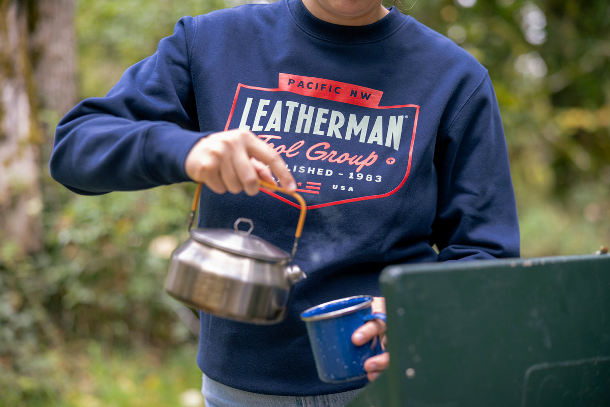 Translation missing: es.Close-up of a person wearing a navy blue Leatherman PNW Crewneck sweatshirt while pouring hot water from a kettle into a blue enamel mug at a campsite