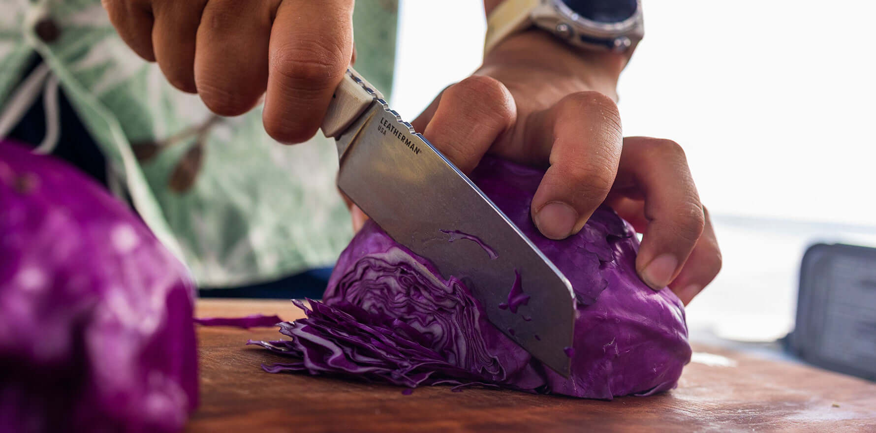 Person using a Leatherman Rustle knife to slice a head of purple cabbage on a wooden cutting board, shown in a close-up with focus on the blade and hands in action.