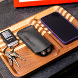 Translation missing: de.A black Leatherman Hypalon tool sleeve holding a multi-tool, neatly placed on a wooden valet tray beside a set of keys and a smartphone with a purple case, showcasing an organized everyday carry setup