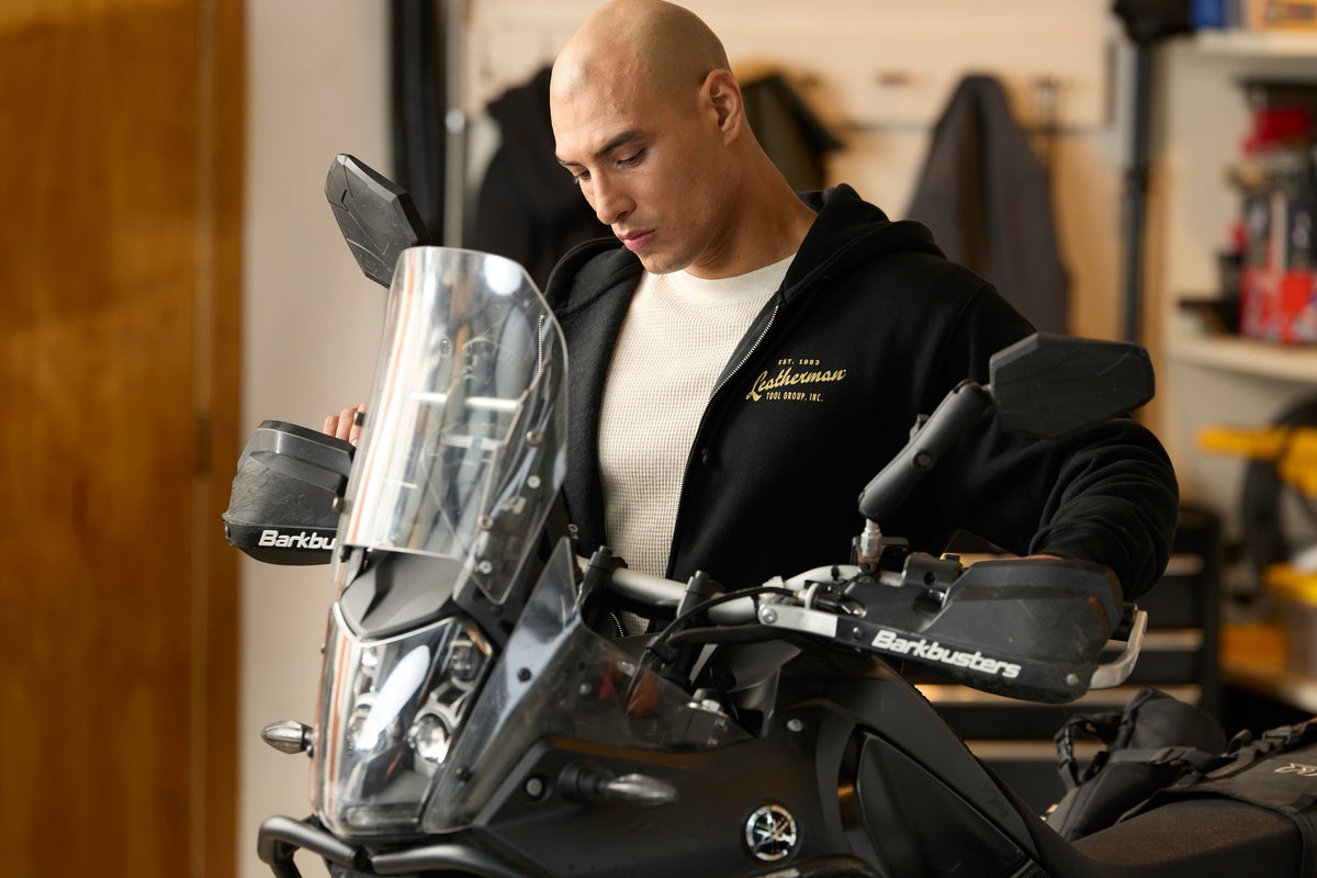 A person wearing a black Leatherman Vintage Zip-Up hoodie standing next to a motorcycle in a garage, with the hoodie’s gold Leatherman logo visible on the chest.