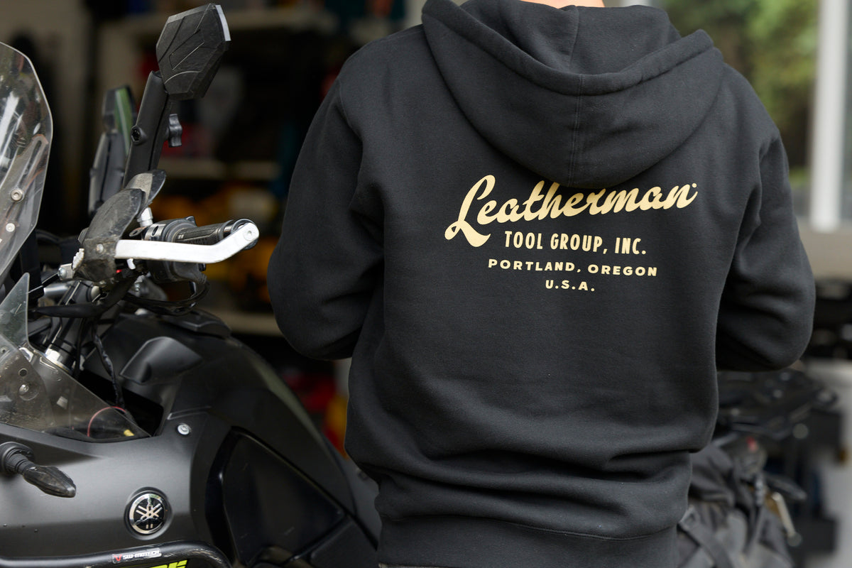 A person wearing a black Leatherman Vintage Zip-Up Hoodie standing next to a motorcycle, showing the gold Leatherman Tool Group, Inc. logo printed on the back.