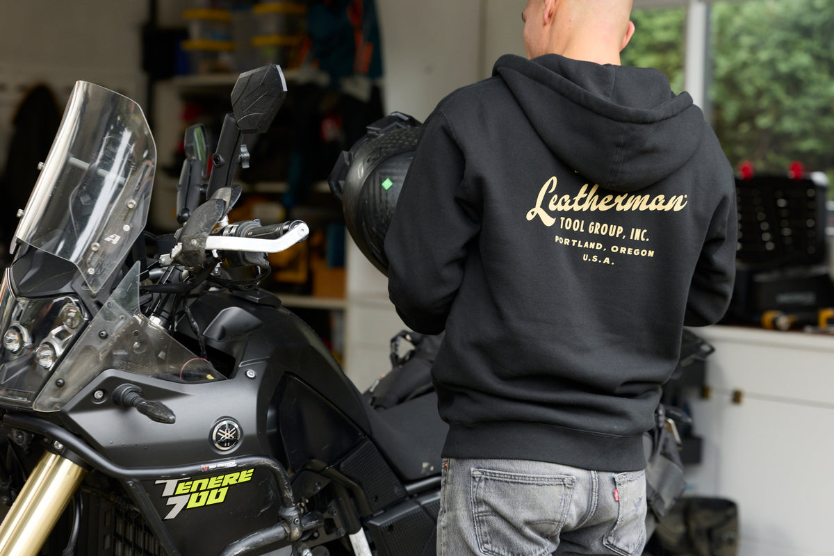 A person wearing a black Leatherman Vintage Zip-Up Hoodie standing next to a motorcycle, showing the gold Leatherman Tool Group, Inc. logo printed on the back.