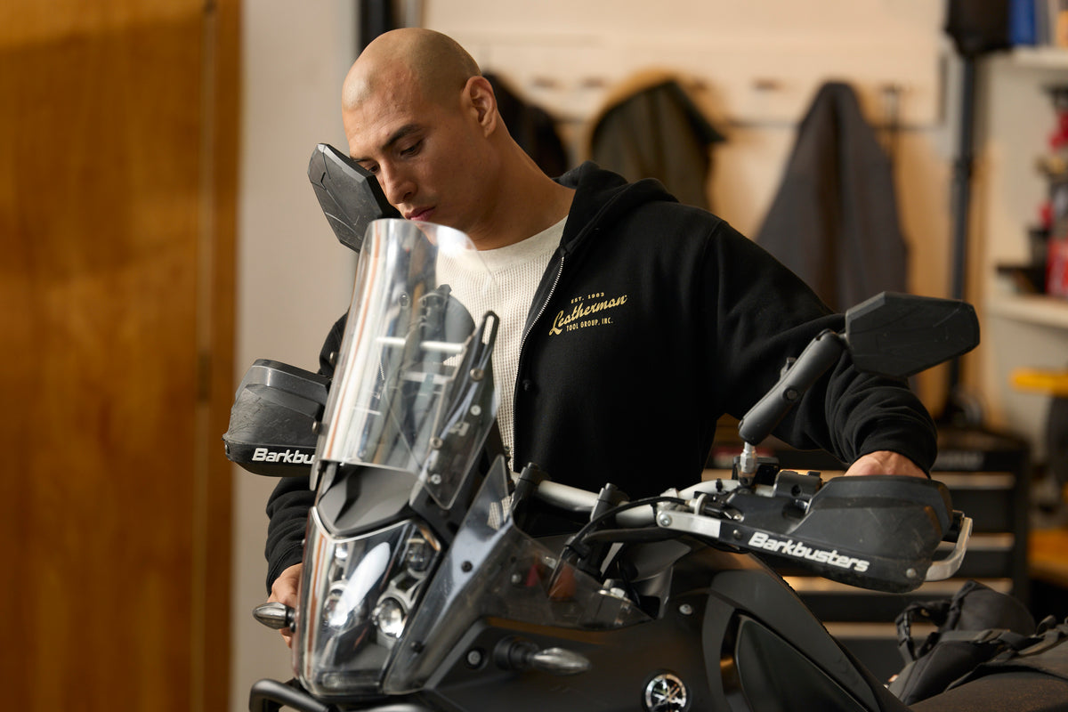 A person wearing a black Leatherman Vintage Zip-Up hoodie standing next to a motorcycle in a garage, with the hoodie’s gold Leatherman logo visible on the chest.